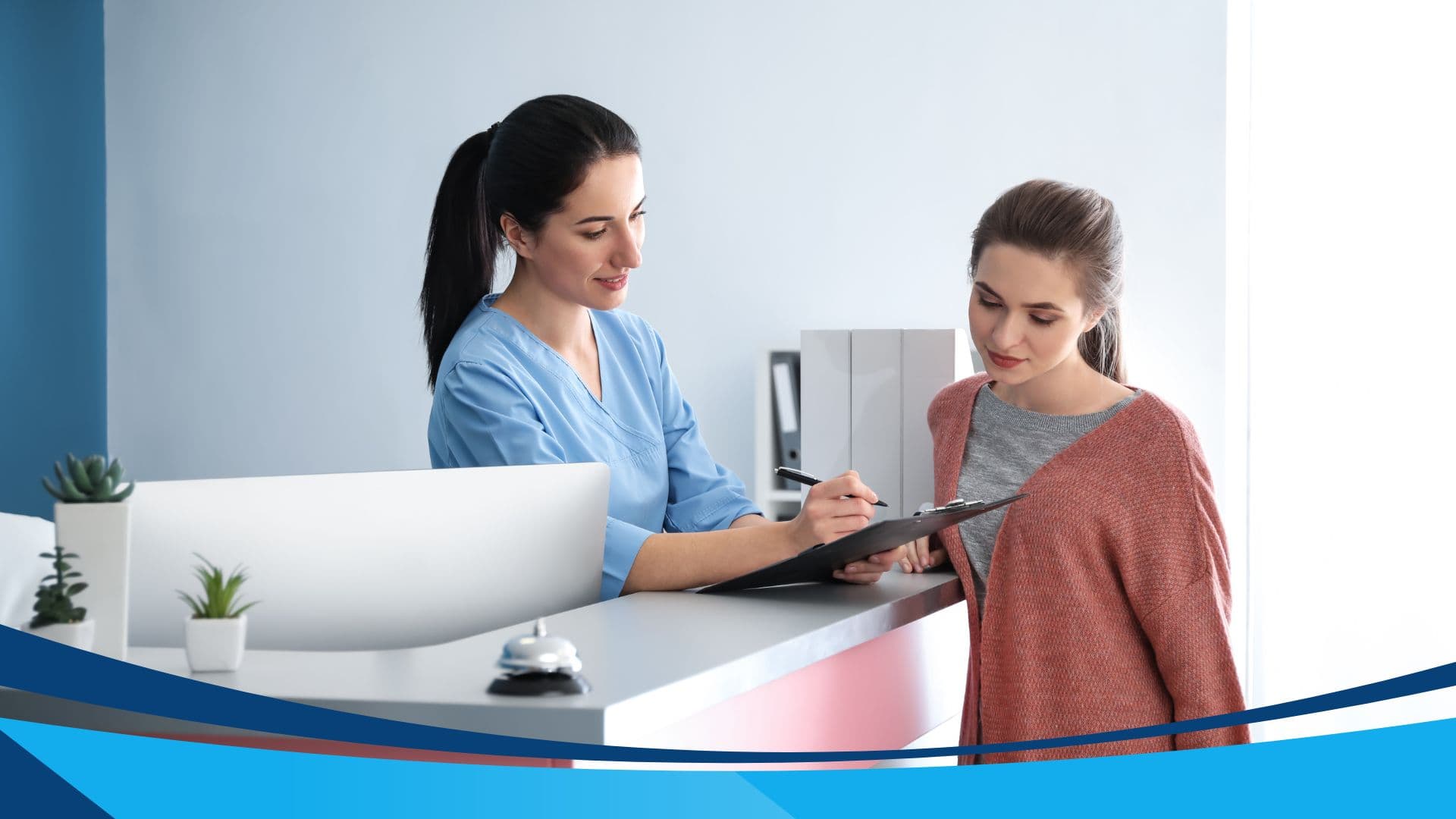 A woman in medical scrubs stands behind a reception desk, writing on a clipboard held by a woman standing in front of the desk. The setting appears to be a clinic or doctor's office with plants and office supplies on the desk.