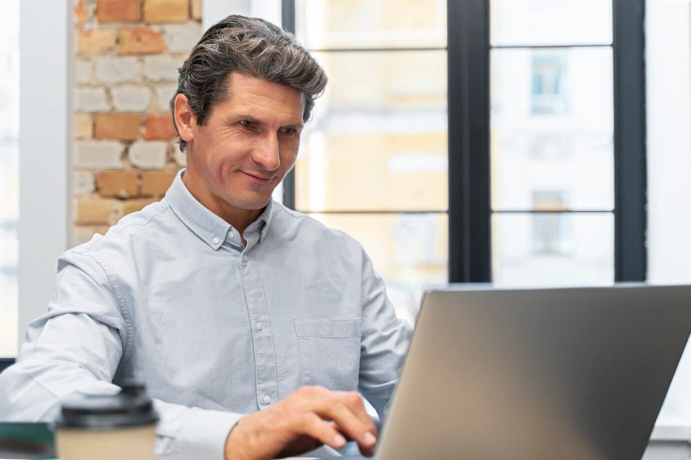 A middle-aged man with salt-and-pepper hair sits at a desk, working on a laptop. He wears a light blue button-down shirt. A brick wall and a multi-paned window appear in the background, and a coffee cup sits next to the laptop.