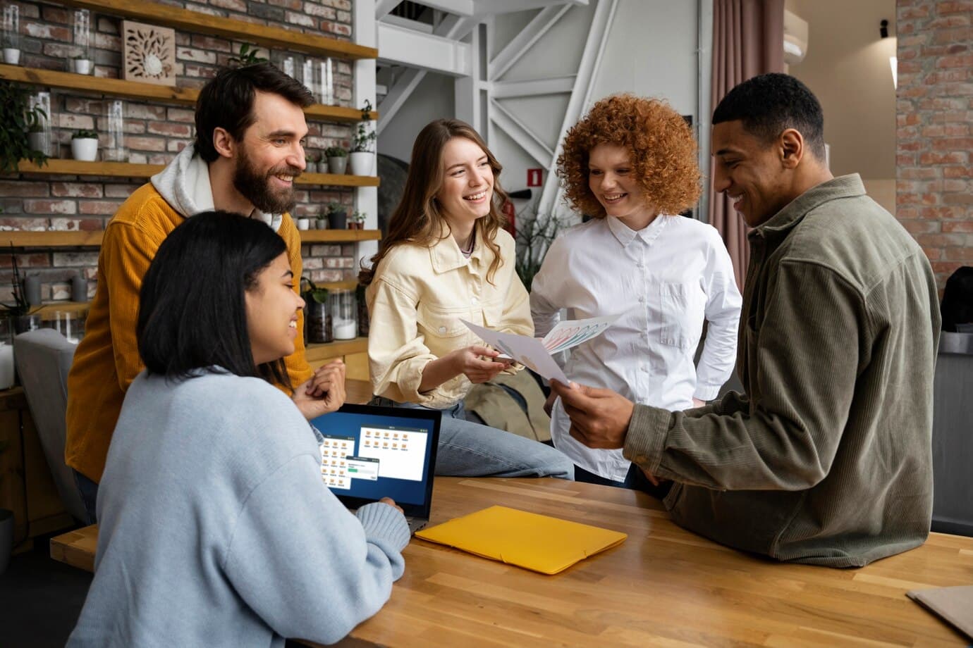A group of five friends in a casual setting share a joyful moment as they gather around a desk, with one person taking a selfie. They are smiling and posing for the camera.