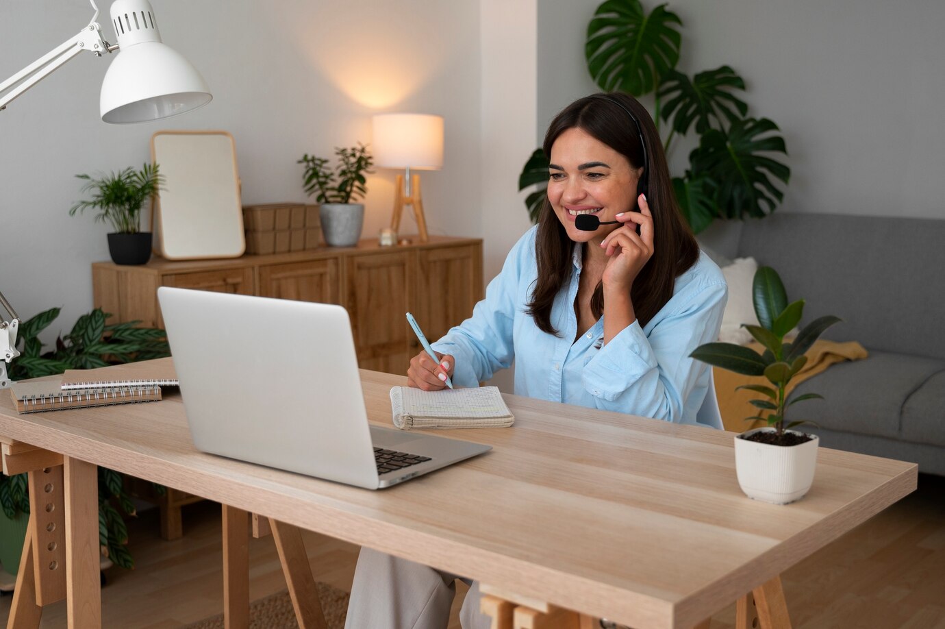 A woman works from home at a light wood desk with a laptop, headset, and notebook; there are plants and a lamp in the background.