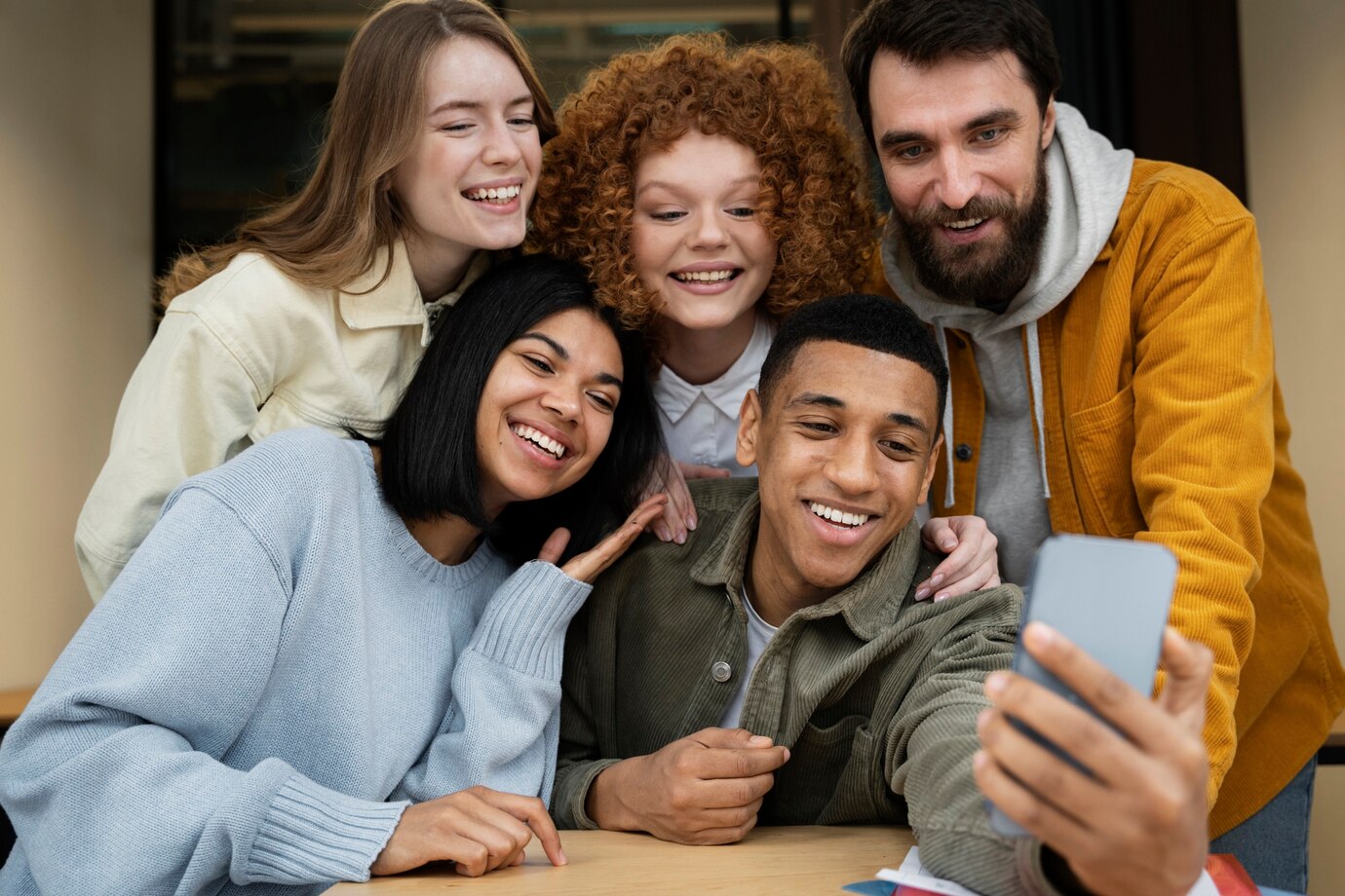 Five friends indoors gather around a table to take a group selfie. They are smiling and laughing, showing a diverse mix of ages and backgrounds, with one person holding the phone to snap the photo.